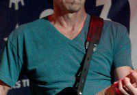 A man playing an electric guitar on stage, wearing glasses and a green shirt.